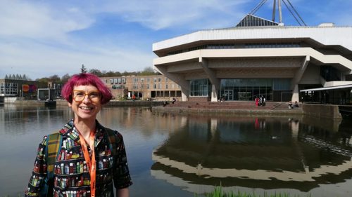 A person with short pink hair, wearing large orange glasses and a dress with a colourful book print, is standing in front of a lake in front of a modernist concrete university building on a clear day