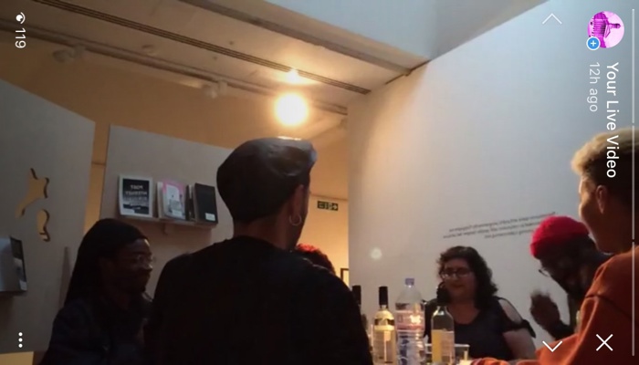 A small group of people gathered around a table in a gallery space, talking beside bottles and books under warm indoor lighting, with a smartphone interface overlay visible on the image.