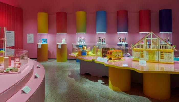 A wide exhibition room with brightly coloured cylindrical columns along the back wall. Several display tables hold Barbie dollhouses and playsets, lit under soft pink lighting.