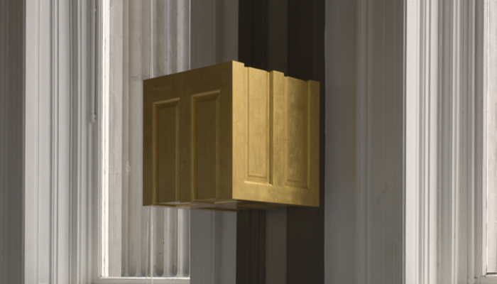A gold, box-like sculptural form made out of a cut up door is mounted to a dark column in a bright gallery space, set against tall windows and a bold black-and-white patterned floor.