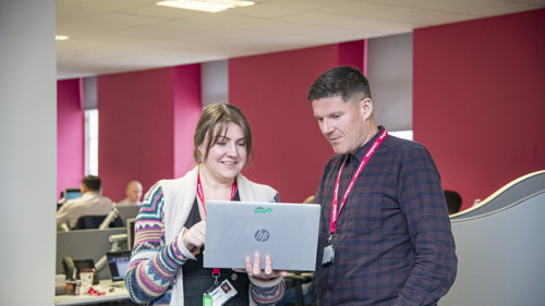 Two people standing and looking at a laptop in an open plan office