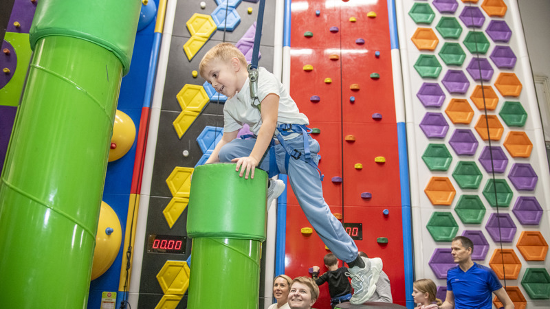 A young boy with a harness on climbing up bright green cylinders with colourful climbing walls in the background.