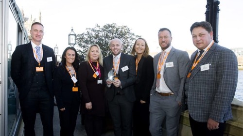 A group of seven people, all wearing smart clothing and orange lanyards, gathered for a group photo on an outdoor balcony area. One of them is holding a glass award