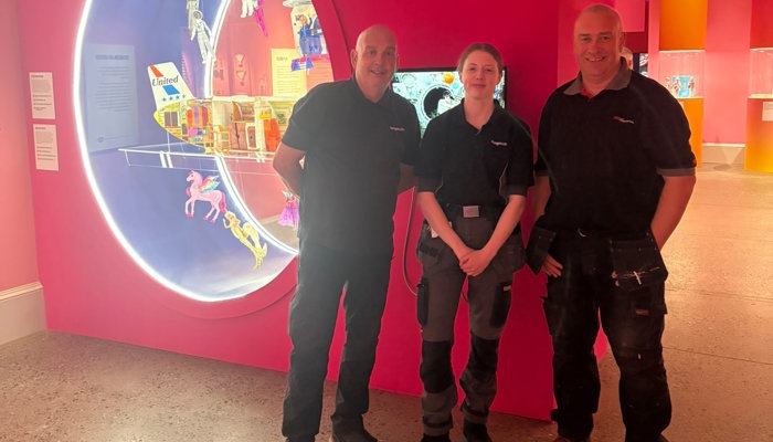 Three people wearing Glasgow Life uniforms are standing and smiling in a large pink exhibition space. There is a pot of pink paint and a roller at their feet