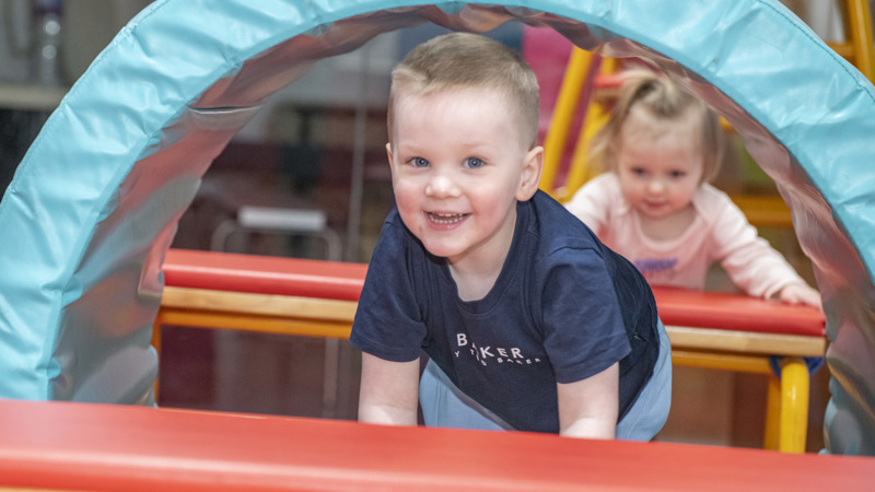 A young child crawls through a soft, padded tunnel structure in an indoor play area, with another child following behind. Brightly coloured mats and play equipment surround them as they move through the obstacle course.