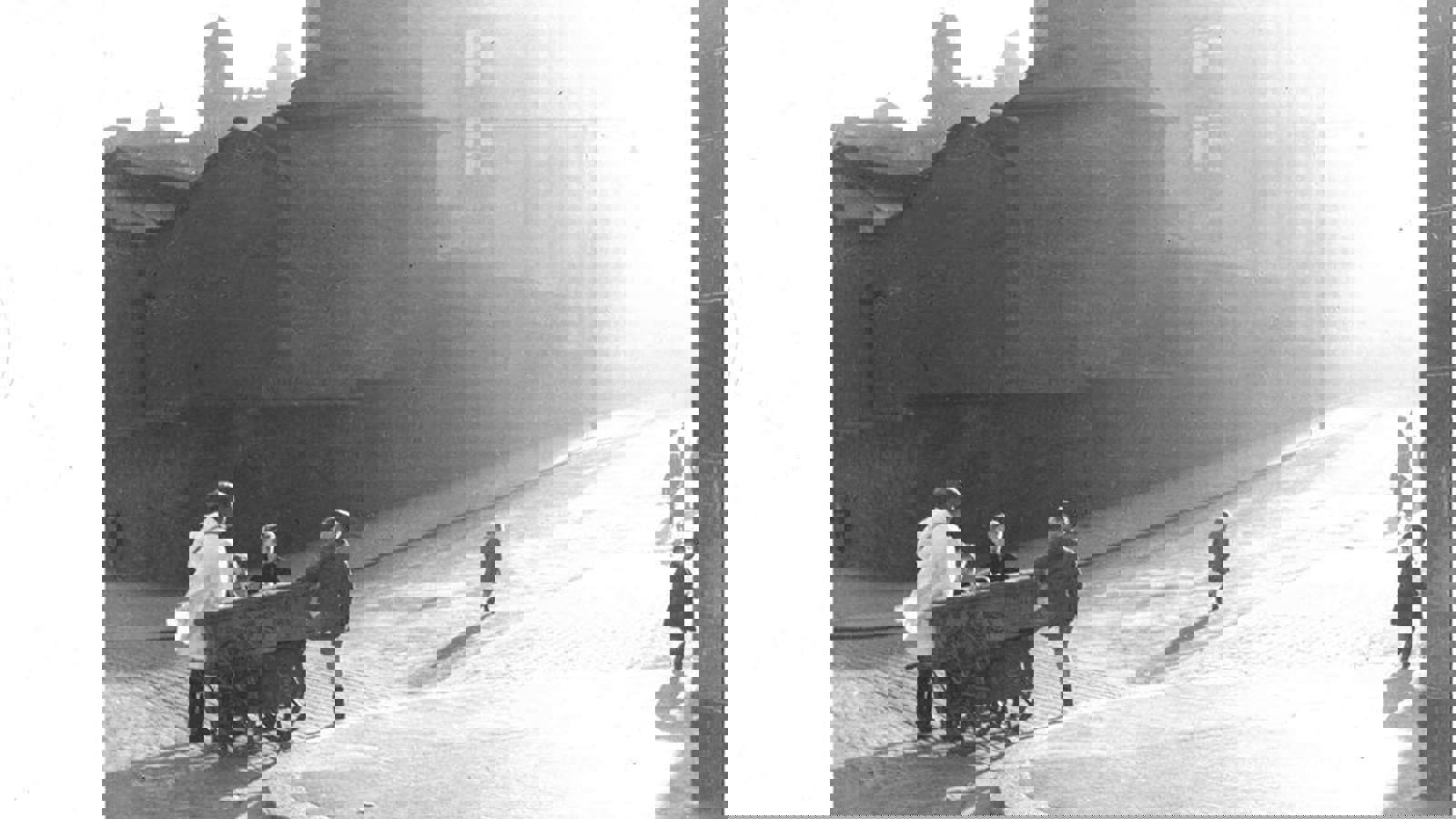 A black and white photograph of young kids playing on sunny cobbled street, some of which are chatting to an adult who is pulling along a small cart.